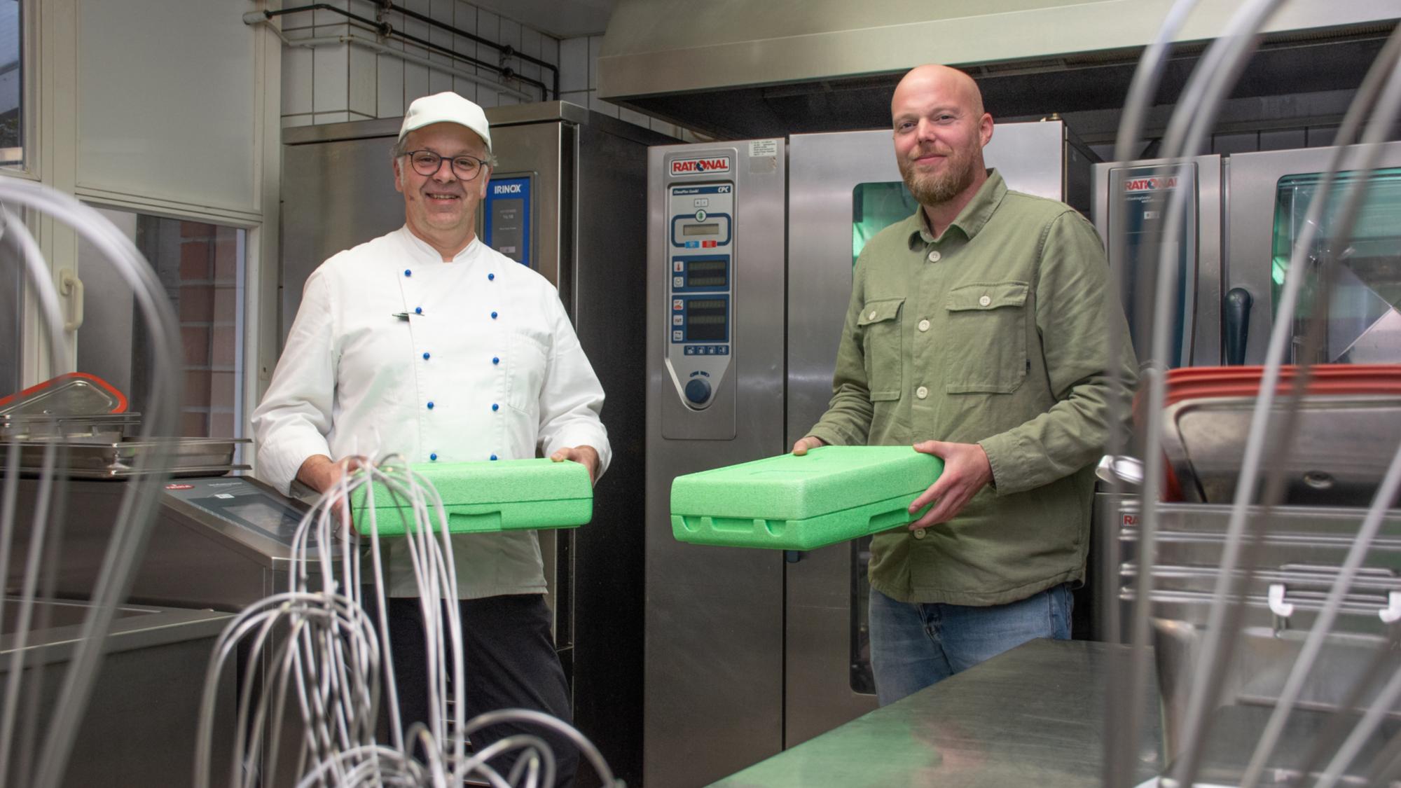 Küchenleiter Markus Lütjann und Einrichtungsleiter Sebastian Stödtke stehen in der Großküche des Henricus-Stifts und zeigen Warmhaltebehälter für das Essen auf Rädern.